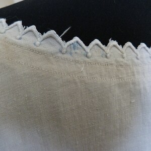 May include: Close-up of a white fabric with a decorative scalloped edge. The edge features a series of inverted V-shapes with small, round accents. The fabric has a textured appearance and visible stitching.