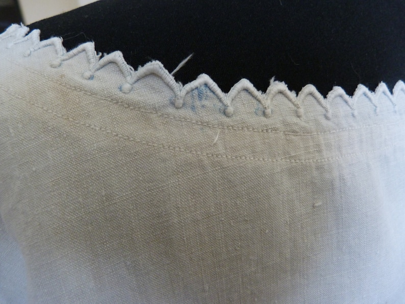 May include: Close-up of a white fabric with a decorative scalloped edge. The edge features a series of inverted V-shapes with small, round accents. The fabric has a textured appearance and visible stitching.