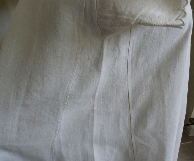 May include: Close-up of a white garment, possibly linen or cotton, with visible seams and a textured surface. The fabric appears to be slightly wrinkled, suggesting a natural fiber. The garment has a collar and a sleeve.