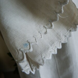 May include: Close-up of off-white fabric with a scalloped edge. The edge is embroidered with a wavy pattern. A small blue stain is visible on the fabric. The fabric appears to be linen or a similar natural material.