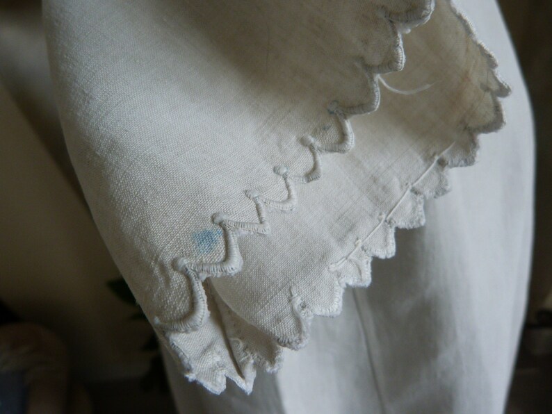 May include: Close-up of off-white fabric with a scalloped edge. The edge is embroidered with a wavy pattern. A small blue stain is visible on the fabric. The fabric appears to be linen or a similar natural material.