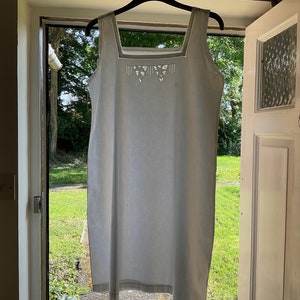 Peut inclure: Une robe blanche sans manches avec un col délicatement brodé d'un motif géométrique. La robe est suspendue à un cintre noir devant une porte blanche.
