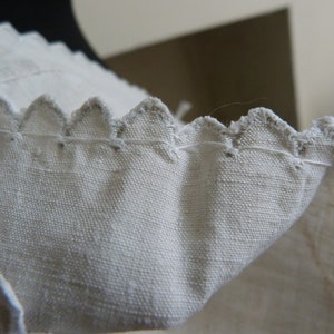 May include: Close-up of a white fabric edge with a scalloped design. The fabric has a textured appearance, and the edges are finished with a contrasting thread. The image highlights the detailed stitching and the delicate, decorative edge.