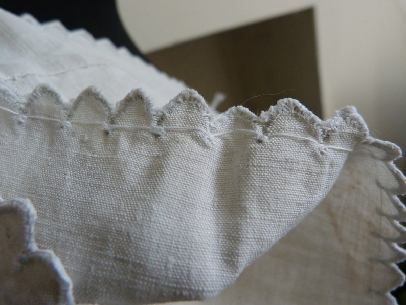 May include: Close-up of a white fabric edge with a scalloped design. The fabric has a textured appearance, and the edges are finished with a contrasting thread. The image highlights the detailed stitching and the delicate, decorative edge.