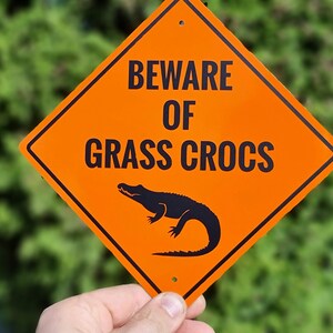 May include: Orange diamond-shaped warning sign with black text that reads "Beware of Grass Crocs". A black silhouette of an alligator is pictured below the text.
