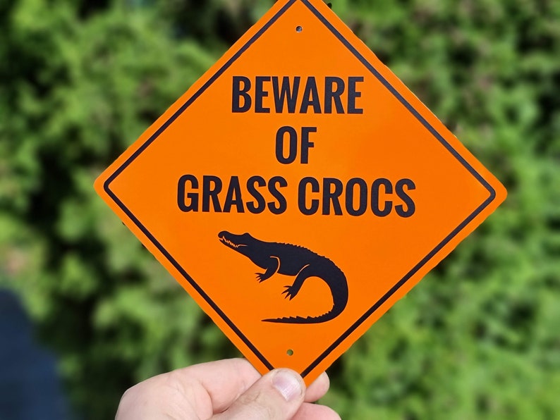 May include: Orange diamond-shaped warning sign with black text that reads "Beware of Grass Crocs". A black silhouette of an alligator is pictured below the text.