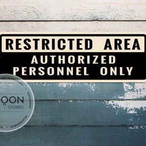 Può includere: Un cartello rettangolare in bianco e nero con la scritta "RESTRICTED AREA AUTHORIZED PERSONNEL ONLY". Il cartello è montato su una superficie in legno blu e bianca, usurata. Un logo circolare con la scritta "BOON STORIES" si trova nell'angolo in basso a sinistra.