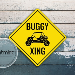 Peut inclure: Un panneau jaune en forme de losange avec une bordure noire présentant une silhouette noire de buggy et le texte BUGGY XING. Un sceau gris circulaire avec le texte printmint et premium grade outdoor sign est placé en bas à gauche.