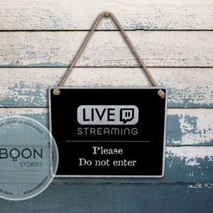Può includere: Un cartello nero con testo argentato che recita "LIVE STREAMING" con il logo di Twitch. Il cartello è appeso su uno sfondo di legno blu e bianco invecchiato. Il testo sotto il logo recita "Si prega di non entrare".