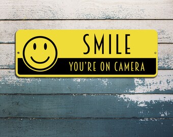 Pode incluir: Um sinal amarelo e preto com um rosto sorridente e o texto "SMILE YOU'RE ON CAMERA".