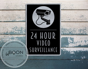 Pode incluir: Uma placa de metal preta e prateada com uma silhueta branca de uma câmera de segurança e o texto "VIGILÂNCIA POR VÍDEO 24 HORAS".