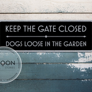 Op de afbeelding: Een rechthoekig zwart metalen bord met witte tekst: "KEEP THE GATE CLOSED" boven "DOGS LOOSE IN THE GARDEN". Het bord is zilverkleurig omlijst en gemonteerd op een verweerde, blauw-witte houten achtergrond.
