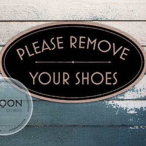 Op de afbeelding: Een zwart ovaal bord met een witte rand en de tekst "PLEASE REMOVE YOUR SHOES" in roze lettertype. Het bord is gemonteerd op een blauwe en witte houten planken achtergrond.