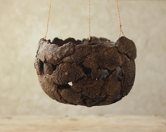 May include: A hanging, textured brown ceramic planter. The planter has a rough, organic appearance with an uneven rim and is suspended by thin, gold-colored wires. The overall shape is a rounded bowl, suitable for plants.