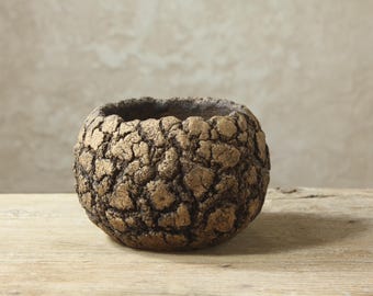 May include: A small, round, bowl-shaped planter featuring a heavily textured, cracked surface with a metallic bronze finish over a dark brown base. The exterior has a rugged, uneven appearance with deep fissures throughout the entire vessel.