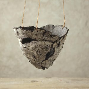 May include: A hanging, handmade ceramic planter with a rough, textured surface. The planter is gray with black accents and is suspended by three thin, golden cords. The planter has an irregular, organic shape, suggesting a natural aesthetic.
