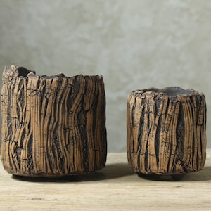 May include: Two cylindrical ceramic planters with a textured, brown, bark-like exterior finish and dark, recessed grooves. The planters feature an uneven, rugged top rim and are supported by small, subtle feet at the base, showcasing a detailed, organic-inspired surface design.