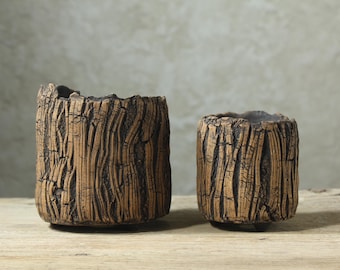 May include: Two cylindrical ceramic planters with a textured, brown, bark-like exterior finish and dark, recessed grooves. The planters feature an uneven, rugged top rim and are supported by small, subtle feet at the base, showcasing a detailed, organic-inspired surface design.
