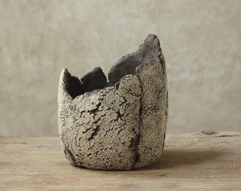 May include: A textured, handmade ceramic pot with a unique, irregular shape. The exterior features a cracked, off-white glaze over a dark base, creating a contrasting, aged appearance. The rim has a jagged, dark edge.