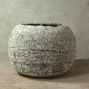 Puede incluir: Un cuenco de cerámica esférico con una superficie muy texturizada y agrietada en tonos blanco roto y gris carbón oscuro. El recipiente presenta una abertura circular ancha en la parte superior y una forma redondeada con una cavidad interior oscura.