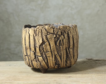 Op de afbeelding: Een cilindervormige keramische plantenpot met een getextureerde, beige buitenkant die lijkt op boomschors. De pot heeft een donkere, onregelmatige rand en rust op drie kleine, donkerbruine voetjes aan de onderkant.