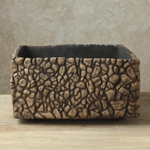 May include: A rectangular planter with a textured exterior surface resembling a collection of small, uneven tan and brown stones. The interior of the container is a solid, dark charcoal gray color, and the overall shape is boxy with straight edges.