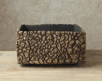 Può includere: Un vaso rettangolare con una superficie esterna strutturata che ricorda una collezione di piccole pietre irregolari color marrone e marrone chiaro. L'interno del contenitore è di un colore grigio antracite scuro e la forma è squadrata.