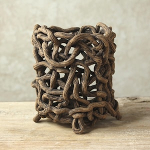 May include: A cylindrical vessel constructed from a dense, interconnected network of brown, textured, root-like forms. The structure features an open-work design with irregular gaps throughout, supported by small, looped extensions at the base.