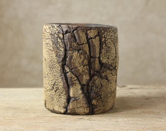 May include: A cylindrical ceramic cup featuring a dark brown base with a heavily textured, cracked surface finish in a metallic gold tone. The exterior displays deep vertical fissures and a rough, uneven appearance across the entire vessel.
