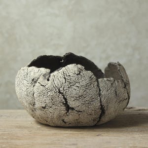 Pode incluir: Uma tigela de cerâmica artesanal com uma superfície texturizada e rachada. A tigela é principalmente esbranquiçada com interior e bordas cinza escuro. Assenta sobre uma superfície de madeira, mostrando sua forma orgânica única.