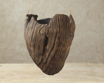 Può includere: Un vaso sospeso in ceramica con un esterno marrone scuro testurizzato che imita l'aspetto della corteccia d'albero. Il contenitore presenta un'apertura profonda e irregolare sulla parte superiore ed è appeso tramite sottili fili metallici dorati.
