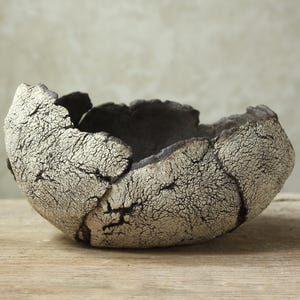 Può includere: Una ciotola in ceramica fatta a mano con una superficie unica e strutturata. La ciotola presenta un esterno screpolato bianco sporco e un interno grigio scuro. La forma organica e la tavolozza dei colori creano un'estetica rustica e artistica. Ideale per l'arredamento.
