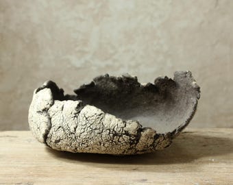 May include: A hand-formed ceramic bowl with a rough, uneven rim and a deep, dark interior. The exterior features a heavily cracked, off-white textured glaze that reveals a dark charcoal base underneath, creating a stark contrast in color.