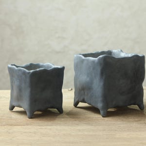 Pode incluir: Dois vasos de cerâmica cinzentos com uma superfície texturizada irregular. Os vasos têm uma forma quadrada com quatro pernas.