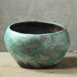 Puede incluir: Un cuenco de cerámica pequeño y redondo con una forma orgánica e irregular. El exterior presenta un esmalte moteado con parches de color turquesa vibrante y verde espuma de mar sobre una base oscura y texturizada. El interior es oscuro.