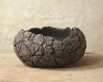 May include: A low, round ceramic bowl with a dark brown, heavily textured exterior that resembles cracked, dry earth. The rim is uneven and organic in shape, while the interior of the bowl is smooth and finished in a deep, matte black glaze.