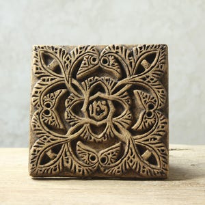 May include: A square, decorative tile with a carved, intricate design. The tile is made of a dark brown material and features a floral pattern with a central motif.