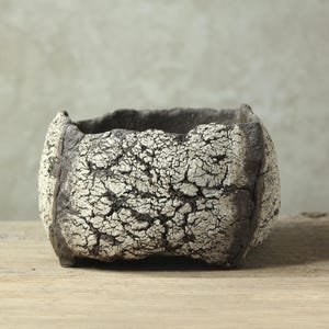 May include: A square, textured ceramic planter with a cracked, off-white surface and dark gray edges. The planter has a rough, organic shape, suitable for small plants or succulents. The neutral color palette complements various decor styles.