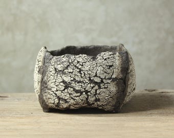 Może przedstawiać: Kwadratowa ceramiczna donica o teksturowanej, kremowej powierzchni i ciemnoszarych krawędziach. Donica ma szorstki, organiczny kształt, odpowiedni dla małych roślin lub sukulentów. Neutralna paleta kolorów uzupełnia różne style wystroju.