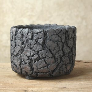 Peut inclure: Un pot de fleurs cylindrique avec un extérieur gris anthracite foncé présentant une surface texturée et profondément craquelée. Le récipient possède un bord épais et irrégulier et repose sur de petits pieds discrets à la base.