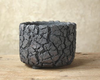 May include: A cylindrical planter with a dark charcoal gray exterior featuring a deeply cracked, textured surface. The vessel has a thick, uneven rim and is supported by small, low-profile feet at the base, creating a rugged and weathered appearance.