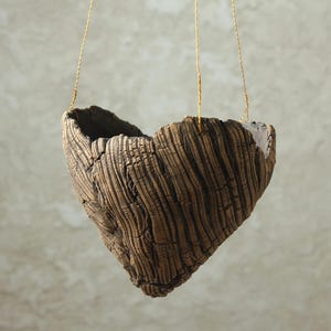 Peut inclure: Un pot de fleurs suspendu en forme de cœur en céramique brune texturée avec une surface profondément rainurée rappelant l'écorce d'arbre. Le récipient est suspendu par de fins fils métalliques de couleur dorée fixés au bord supérieur.