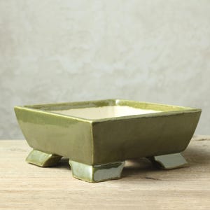 May include: A square green ceramic bonsai pot with four short legs. The pot has a white glazed interior.