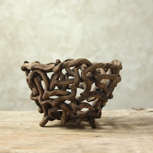 Könnte beinhalten: Eine Schale aus ineinander verschlungenen, dunkelbraunen, knorrigen Ästen. Die Struktur weist ein offenes, gitterartiges Design mit zahlreichen Lücken zwischen den gedrehten Holzelementen auf und bildet eine abgerundete, organische Form.