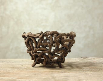 May include: A bowl constructed from interwoven dark brown gnarled branches. The structure features an open, lattice-like design with multiple gaps between the twisted wooden elements, forming a rounded, organic shape with a stable base.