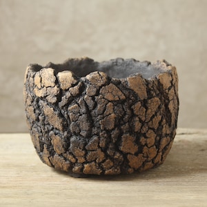 Op de afbeelding: Een ronde, komvormige plantenbak met een sterk getextureerd, gebarsten buitenoppervlak. De afwerking toont een diepe, donkerbruine basiskleur met metallic bronzen accenten op de verhoogde, onregelmatige randen van het gebarsten patroon.