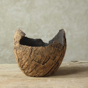 Può includere: Una ciotola in ceramica marrone strutturata, con una forma organica unica. La ciotola ha un esterno ruvido, simile alla corteccia, e un interno scuro. Il bordo superiore è irregolare, aggiungendo fascino rustico. Perfetta per l'arredamento.