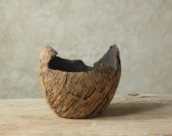 Może przedstawiać: Ręcznie wykonana, teksturowana, brązowa ceramiczna miska o unikalnym, organicznym kształcie. Miska ma szorstką, przypominającą korę zewnętrzną powierzchnię i ciemne wnętrze. Nierówna górna krawędź dodaje rustykalnego uroku.