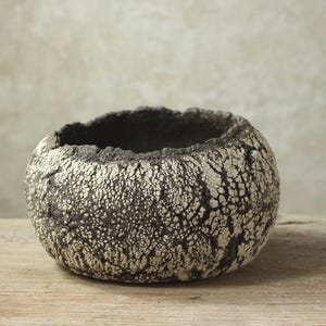 May include: A textured, round ceramic bowl with a dark interior and a rough, cracked exterior. The bowl's surface is a mix of dark brown and off-white, creating a unique, organic appearance. The rim has a jagged edge. The bowl sits on a wooden surface.