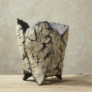 May include: A ceramic vessel with a textured, crackled beige and black exterior finish. The pot features an irregular, jagged rim and stands on three short, pointed feet. The surface displays a deep, dark contrast between the light cracked outer layer and the dark base material visible within the fissures.